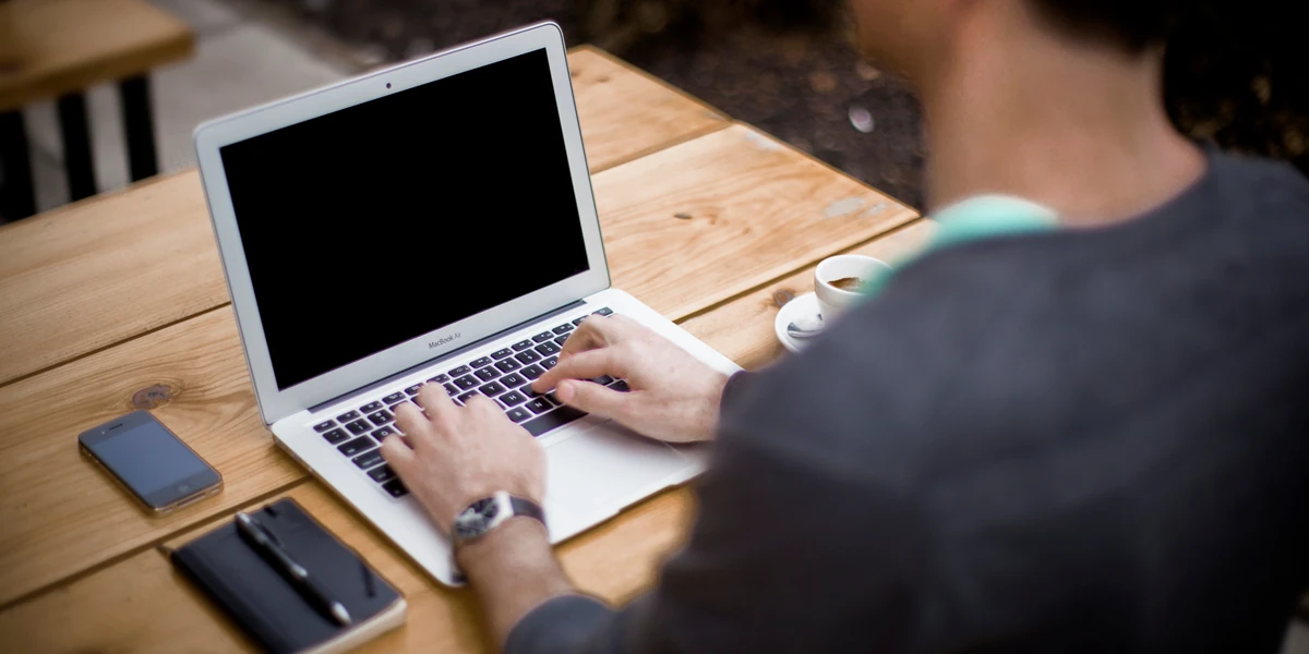 A person seen from behind typing on a laptop at a sunlit wooden table, with an espresso cup, a phone and a notebook beside them